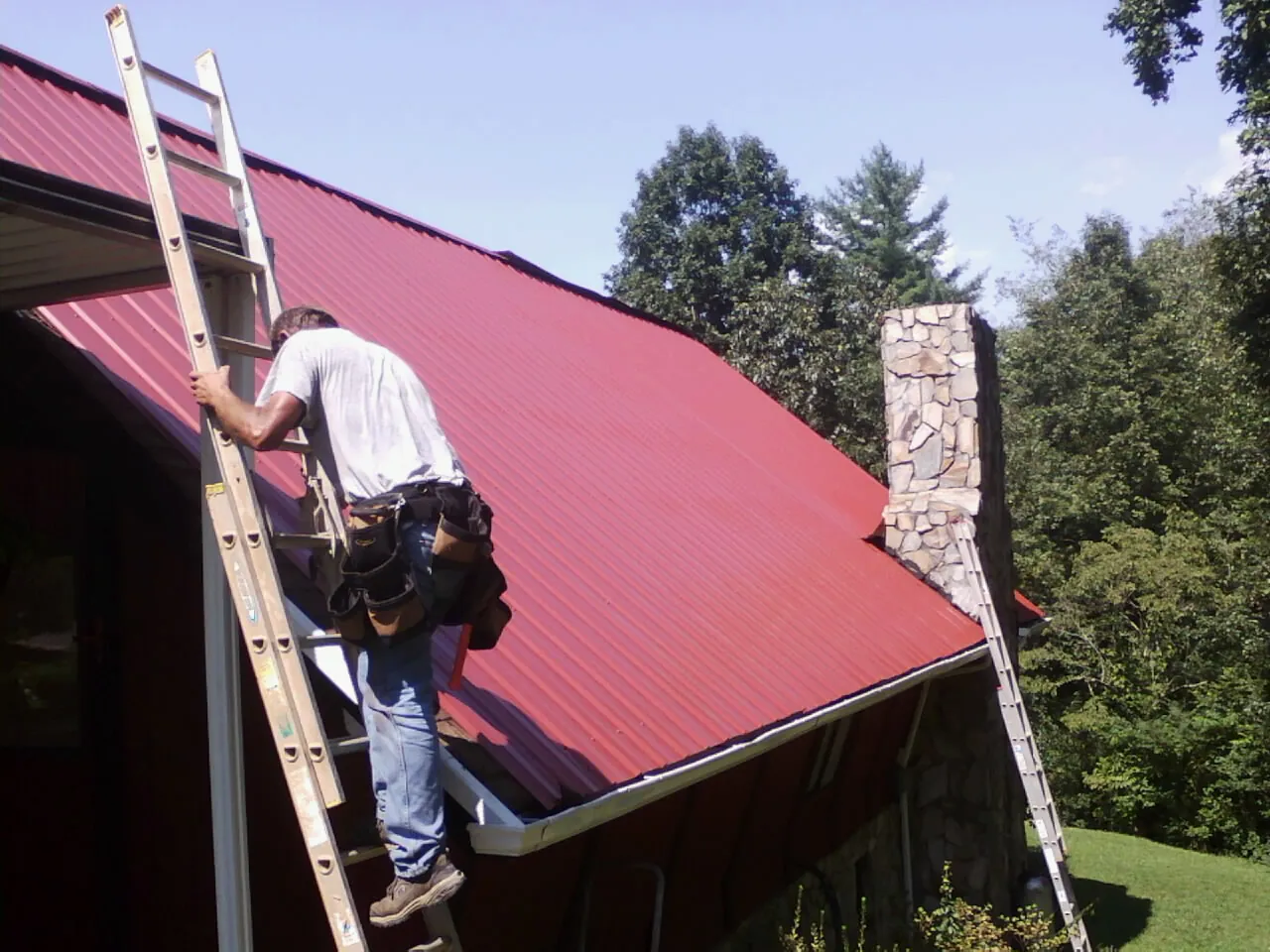 Roofer on a ladder beside a red metal roof and stone chimney — Soffit & Fascia Repair by Roofing Brothers in Duncanville
